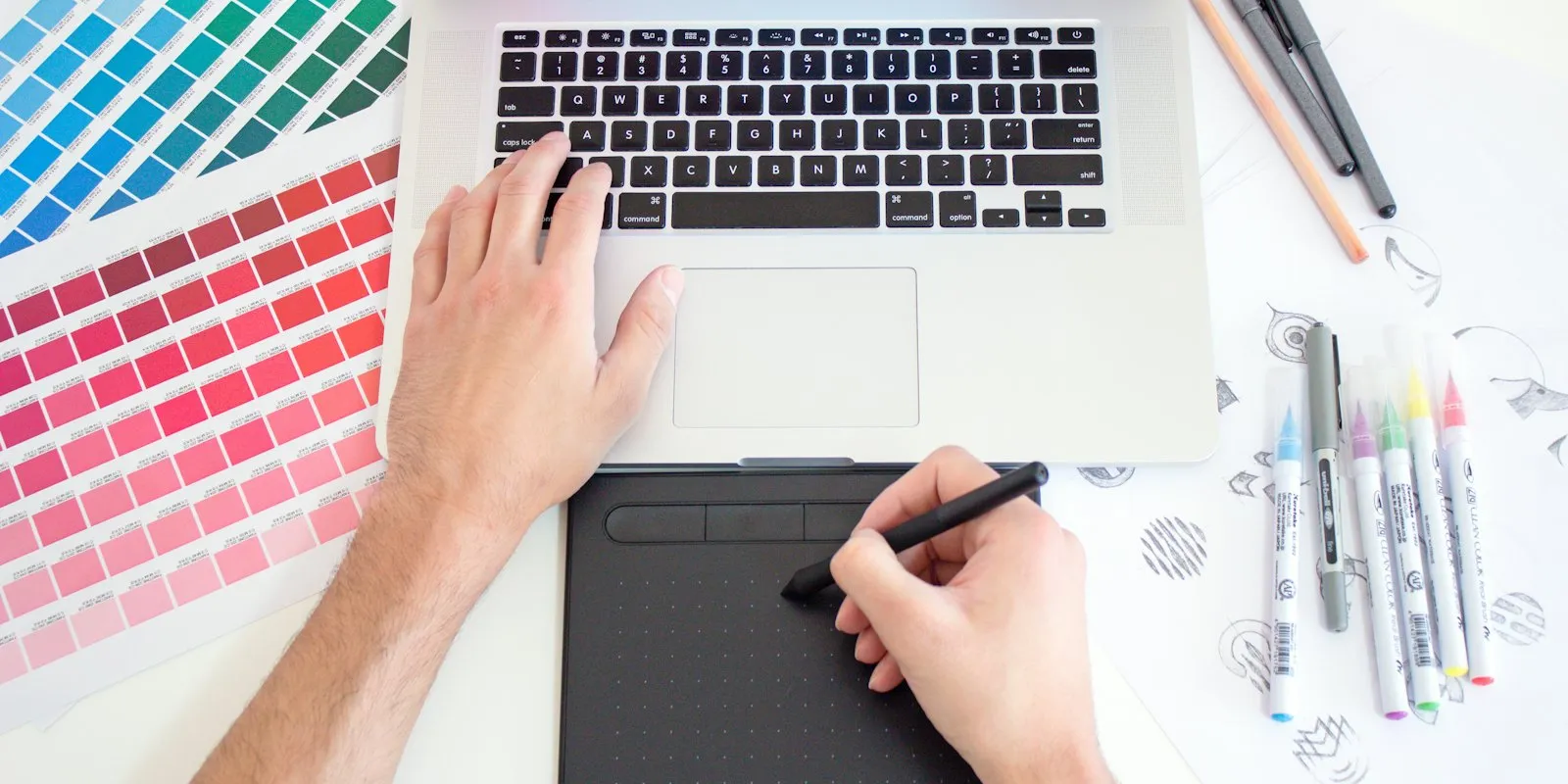 Une main tenant un stylet sur une tablette graphique, posée à côté d'un clavier d'ordinateur portable et de feutres de dessin
